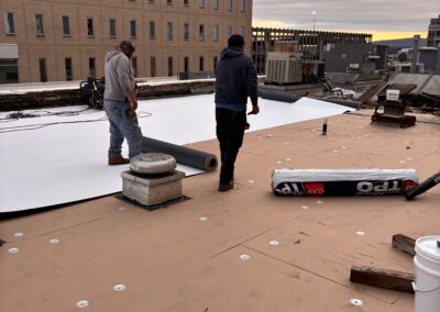 Two men are installing a large roll of plastic on a roof, demonstrating teamwork and construction skills.